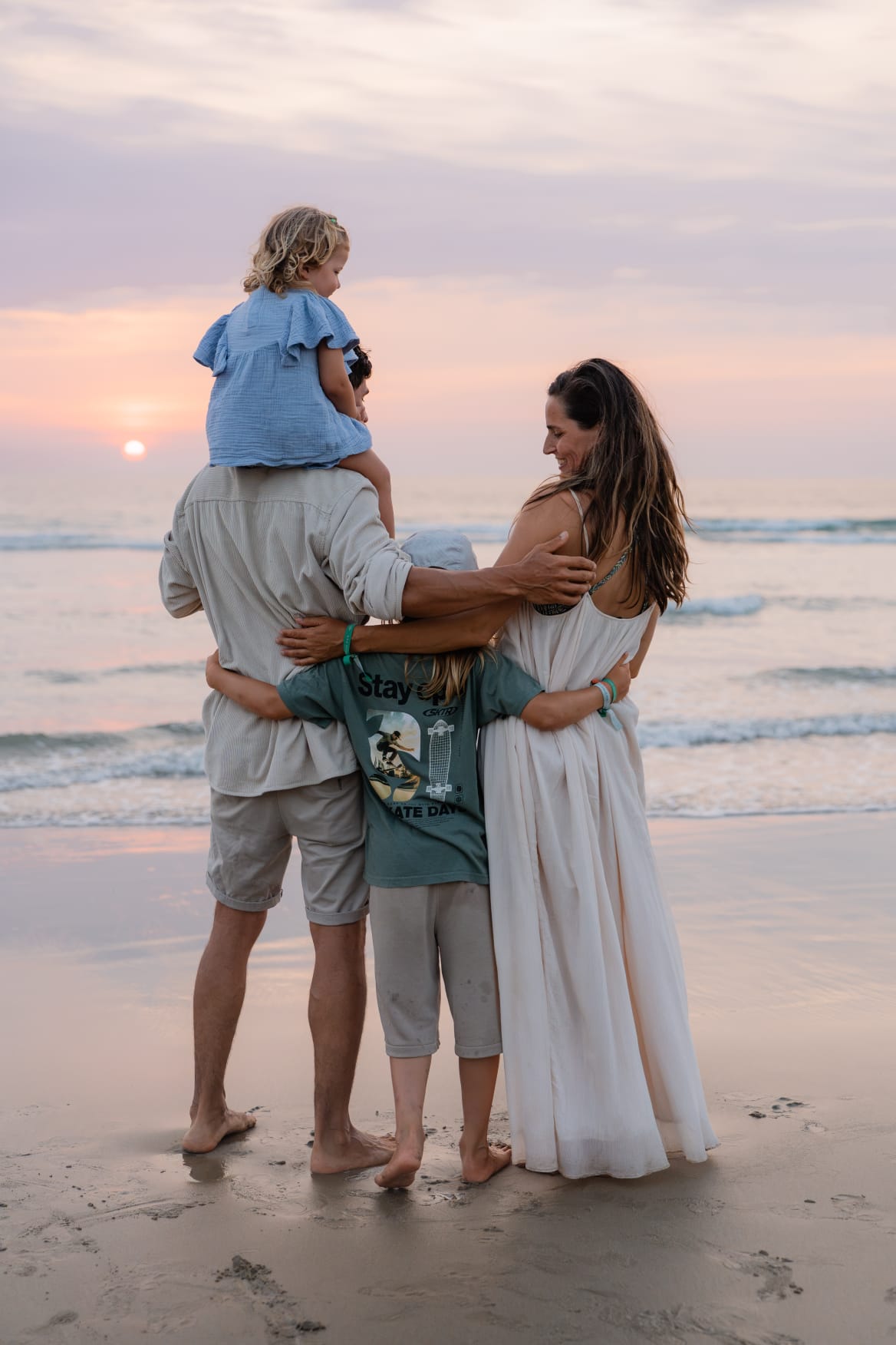 Gezin staat samen aan zee bij zonsondergang, de vader draagt een kind op de schouders